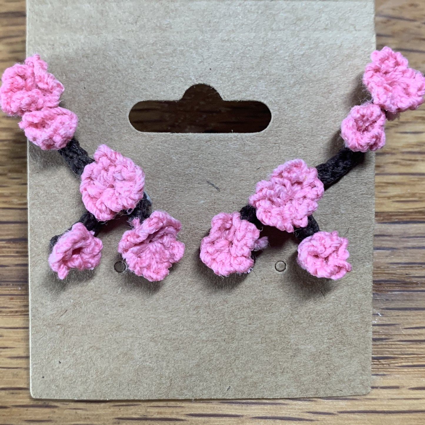 A close-up of stud earrings on an earring card, which is laying on a wooden table. Each earring is shaped like a tiny tree branch, each with six cherry blossom blooms. The branches are designed to "grow" up the lobe towward the helix.